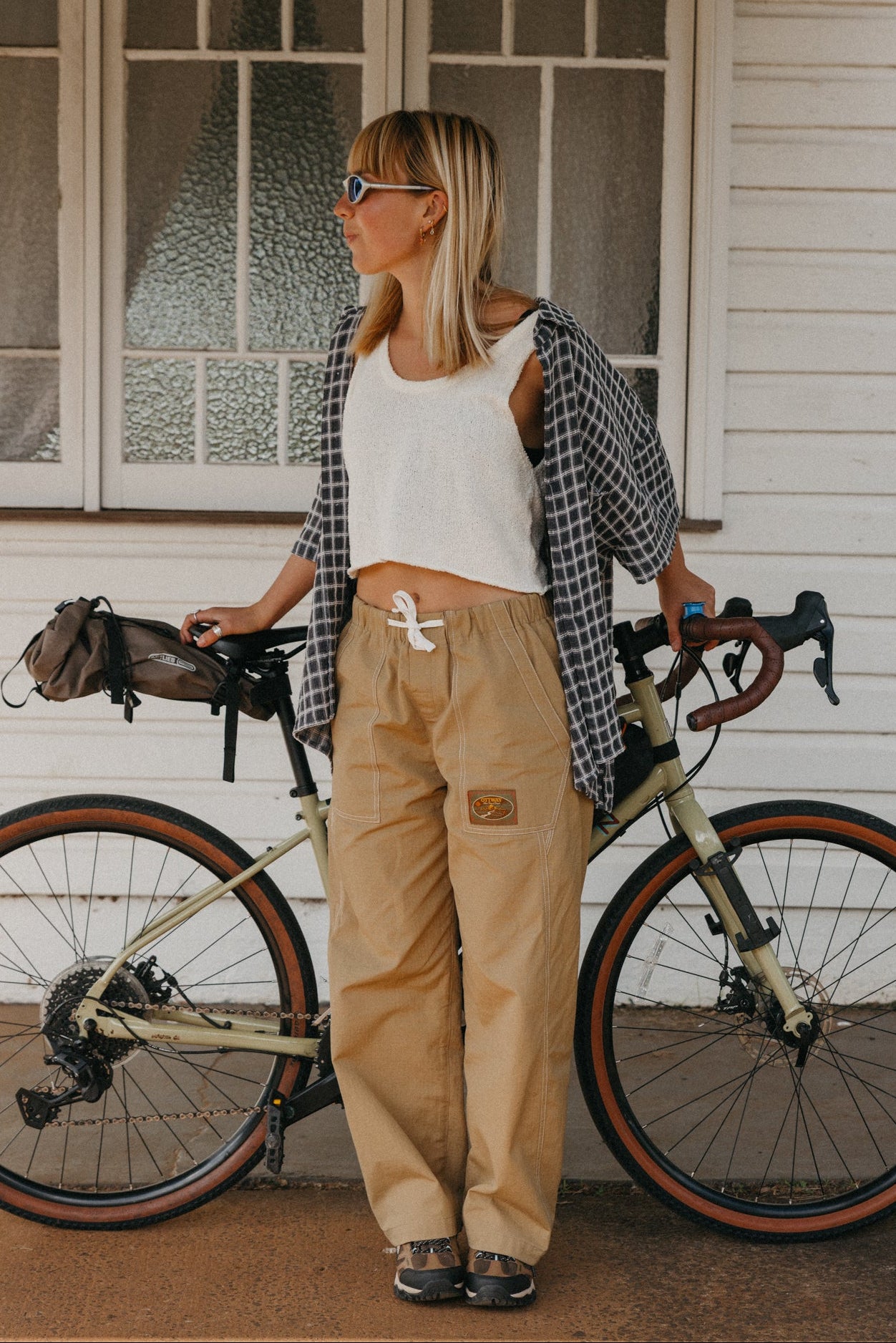A woman stands outdoors next to a bicycle in front of a white house, wearing beige cargo pants, a white crop top, a black-and-white plaid shirt, and sunglasses.