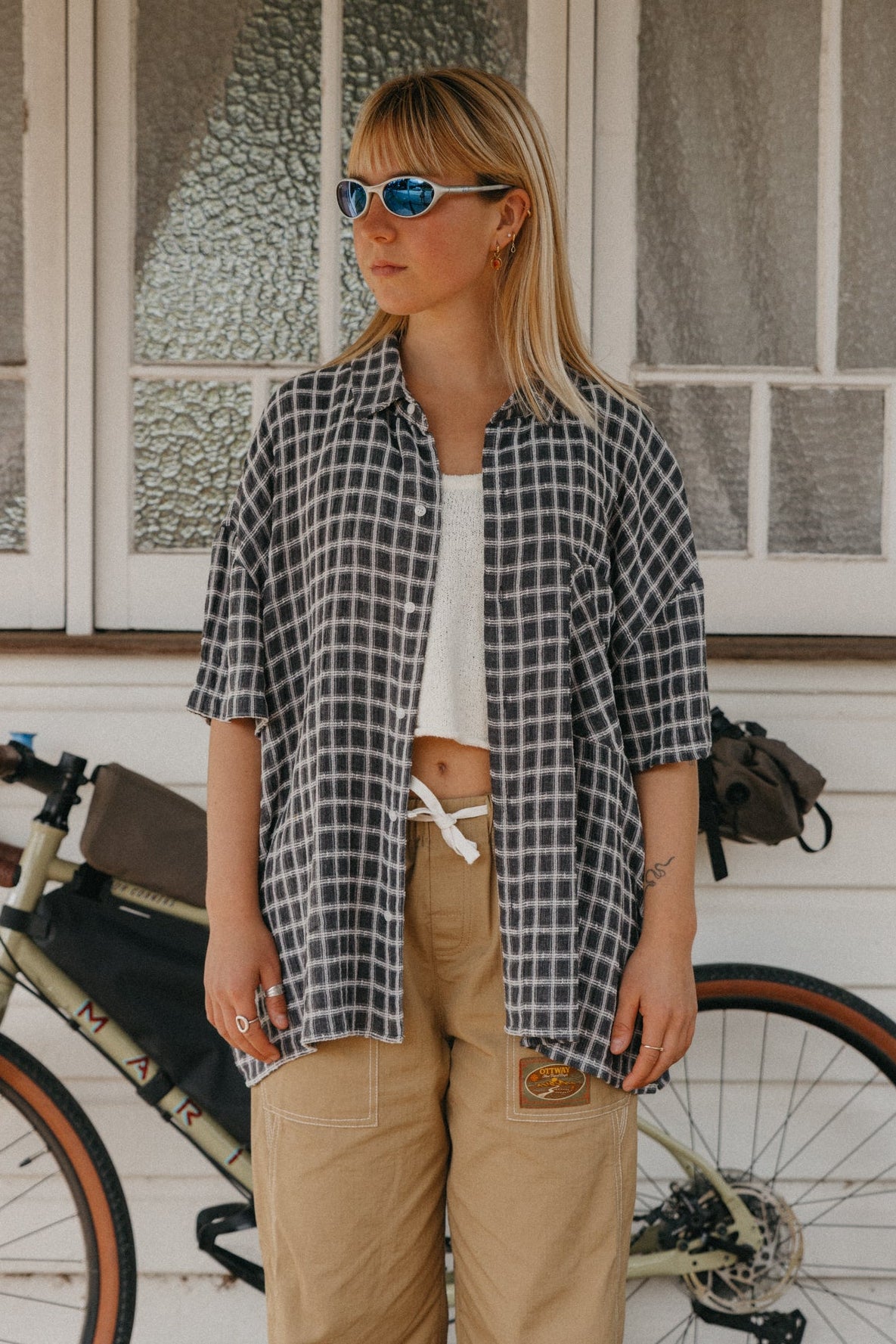 A woman wearing a black and white checkered short-sleeve button-up shirt, tan cargo pants, and sunglasses stands in front of a white house with a bicycle visible behind her.
