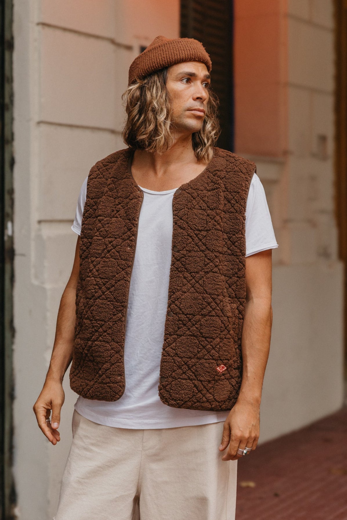 Man wearing a brown quilted vest and beanie standing against a building.