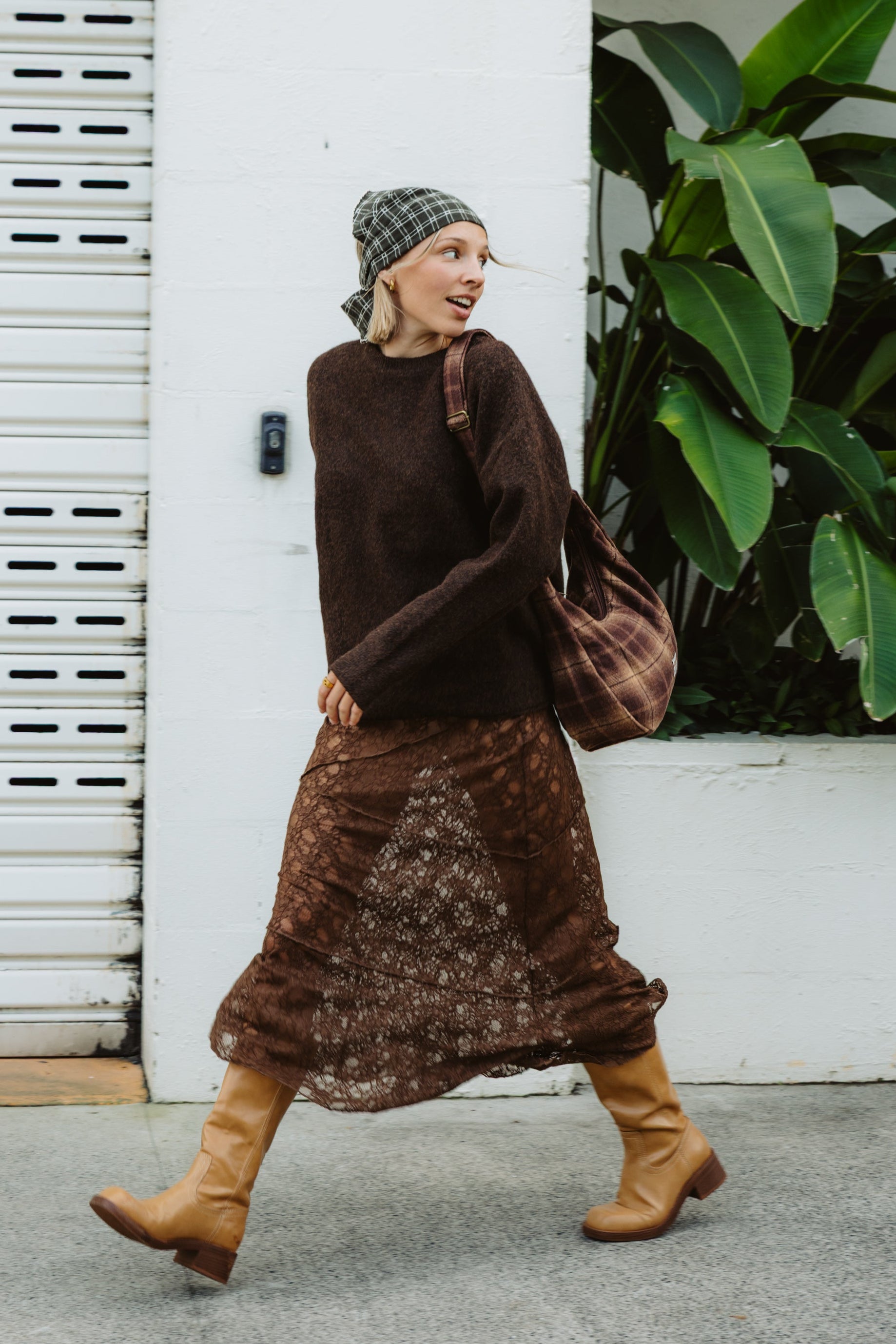 Woman in brown outfit with patterned skirt and hat, standing next to a white wall and plant.